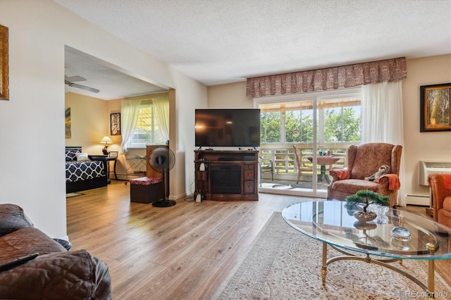 living room featuring ceiling fan, baseboard heating, a wall mounted AC, a textured ceiling, and light wood-type flooring