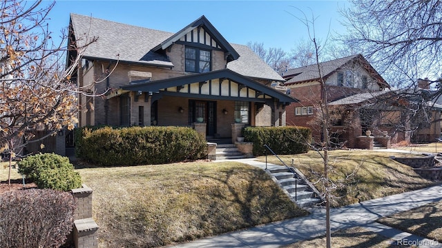 english style home featuring a porch, brick siding, and roof with shingles