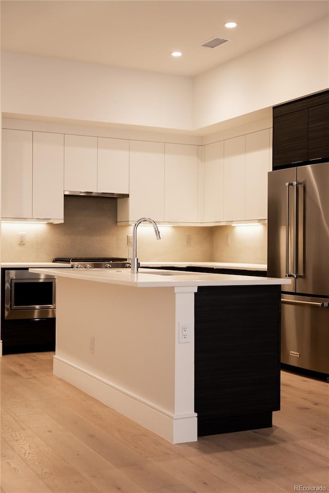 kitchen with appliances with stainless steel finishes, white cabinetry, backsplash, light hardwood / wood-style floors, and an island with sink