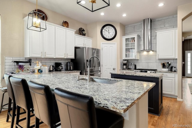 kitchen featuring wall chimney exhaust hood, sink, a center island with sink, pendant lighting, and white cabinets