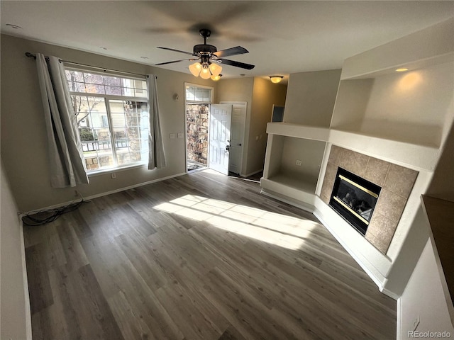 unfurnished living room with ceiling fan, a tiled fireplace, wood finished floors, and baseboards