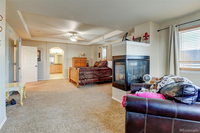 bedroom featuring ceiling fan, ensuite bathroom, a multi sided fireplace, a tray ceiling, and light carpet