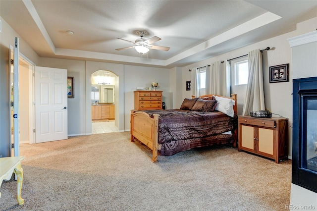 bedroom with a raised ceiling, ensuite bathroom, light carpet, and ceiling fan