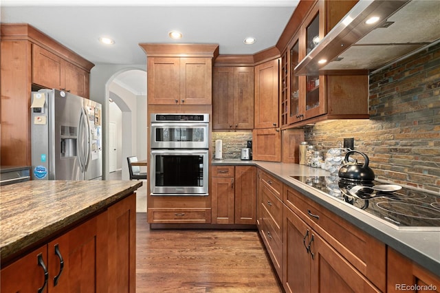 kitchen featuring appliances with stainless steel finishes, wood-type flooring, decorative backsplash, dark stone counters, and wall chimney exhaust hood