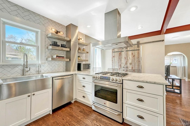kitchen featuring a peninsula, island range hood, arched walkways, a sink, and appliances with stainless steel finishes