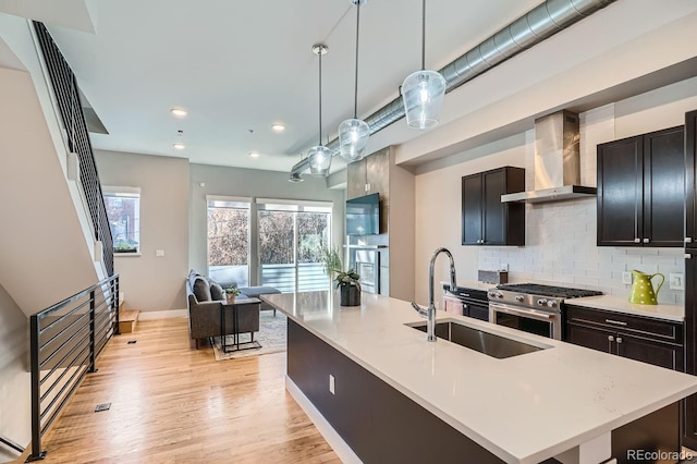 kitchen featuring stainless steel stove, sink, hanging light fixtures, wall chimney range hood, and a center island with sink