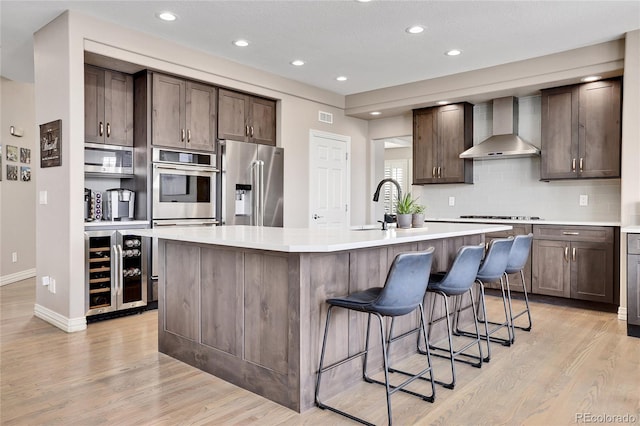 kitchen featuring wall chimney exhaust hood, a breakfast bar, a center island with sink, stainless steel appliances, and beverage cooler