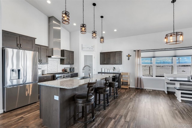 kitchen featuring wall chimney range hood, sink, a kitchen island with sink, stainless steel appliances, and dark brown cabinets