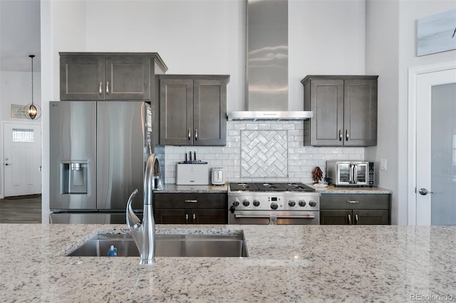 kitchen featuring appliances with stainless steel finishes, wall chimney range hood, sink, hanging light fixtures, and light stone counters