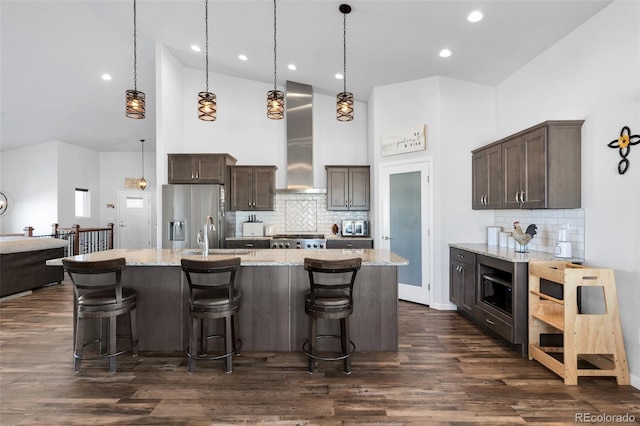 kitchen featuring stainless steel refrigerator with ice dispenser, wall chimney exhaust hood, hanging light fixtures, an island with sink, and light stone countertops