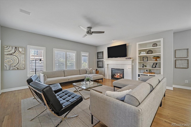 living room with built in shelves, light wood-style flooring, a ceiling fan, baseboards, and a tiled fireplace