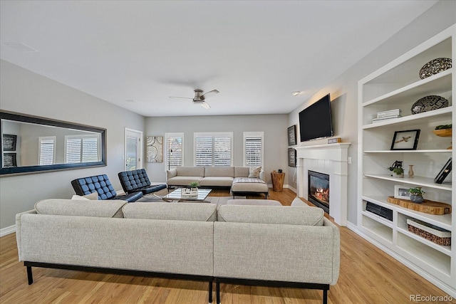 living room featuring built in shelves, a ceiling fan, baseboards, light wood-type flooring, and a glass covered fireplace