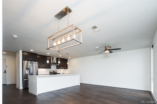 kitchen with dark wood-type flooring, hanging light fixtures, an island with sink, stainless steel appliances, and wall chimney range hood