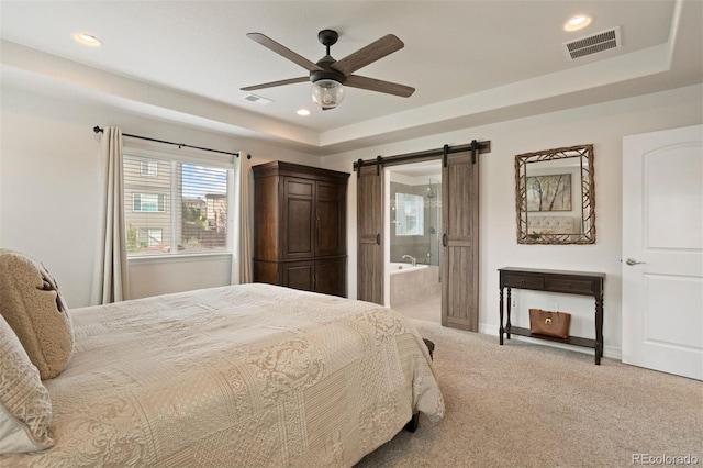 bedroom featuring ceiling fan, a tray ceiling, a barn door, light colored carpet, and ensuite bathroom