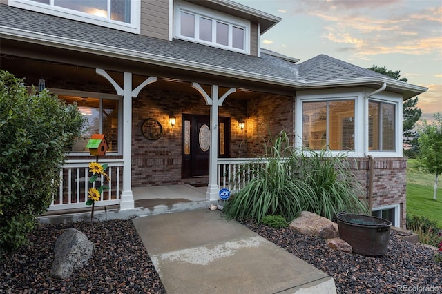 exterior entry at dusk with a porch, brick siding, and roof with shingles