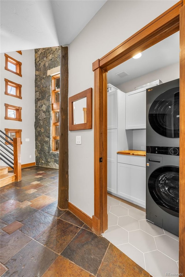 laundry area featuring baseboards, cabinet space, stacked washer and clothes dryer, and stone tile flooring