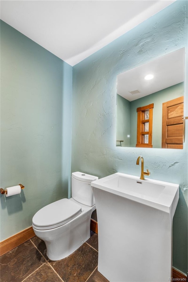 half bathroom with vanity, visible vents, baseboards, toilet, and a textured wall