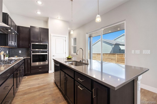 kitchen with appliances with stainless steel finishes, a sink, wall chimney exhaust hood, backsplash, and light wood-style flooring