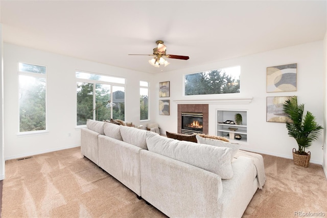 living room with a tiled fireplace, ceiling fan, built in features, and light colored carpet