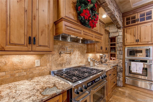 kitchen featuring premium range hood, light hardwood / wood-style flooring, stainless steel appliances, light stone countertops, and decorative backsplash