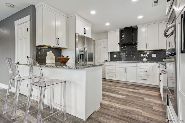 kitchen featuring stone countertops, a peninsula, stainless steel refrigerator with ice dispenser, and wall chimney exhaust hood