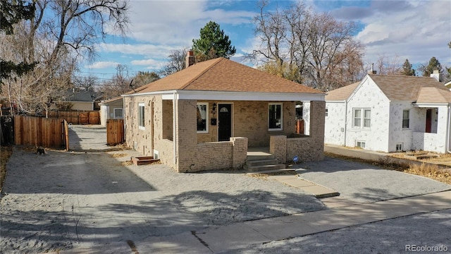 bungalow featuring covered porch, a shingled roof, brick siding, fence, and a chimney