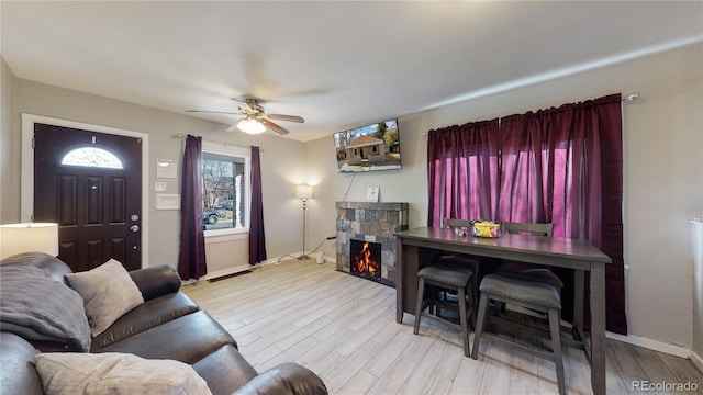 living room featuring a warm lit fireplace, ceiling fan, baseboards, and light wood-style floors