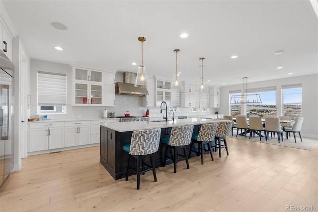 kitchen with wall chimney range hood, white cabinetry, light countertops, light wood-style floors, and an island with sink