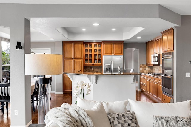 kitchen with brown cabinetry, stainless steel appliances, dark wood finished floors, decorative backsplash, and a tray ceiling
