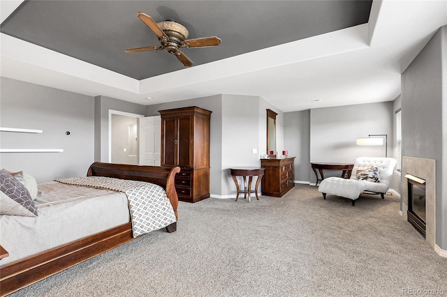 bedroom with ceiling fan, light carpet, a tray ceiling, baseboards, and a glass covered fireplace