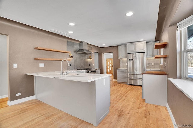 kitchen with wall chimney exhaust hood, tasteful backsplash, light hardwood / wood-style floors, kitchen peninsula, and stainless steel refrigerator