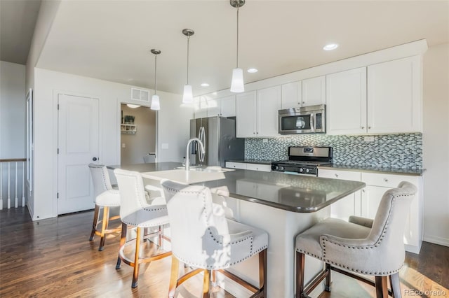 kitchen with dark wood-type flooring, appliances with stainless steel finishes, white cabinetry, hanging light fixtures, and a center island with sink