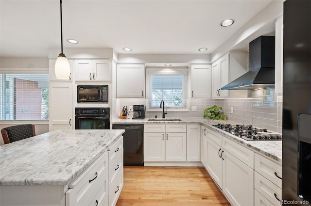 kitchen featuring wall chimney range hood, light wood-style floors, white cabinets, black appliances, and a sink