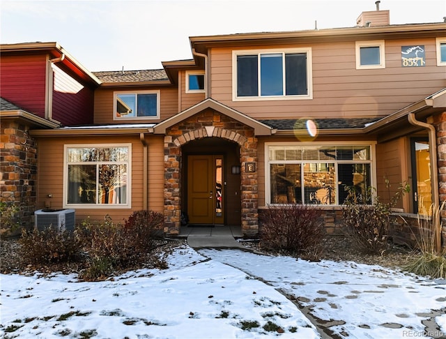 snow covered property entrance featuring stone siding, roof with shingles, a chimney, and central air condition unit