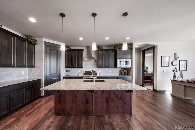 kitchen with decorative light fixtures, wall chimney range hood, a kitchen island with sink, light stone countertops, and stainless steel appliances