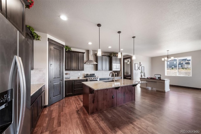 kitchen featuring a breakfast bar area, stainless steel appliances, a center island with sink, decorative light fixtures, and wall chimney exhaust hood