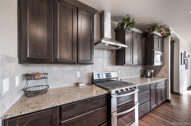 kitchen featuring appliances with stainless steel finishes, dark hardwood / wood-style flooring, light stone counters, dark brown cabinets, and wall chimney exhaust hood