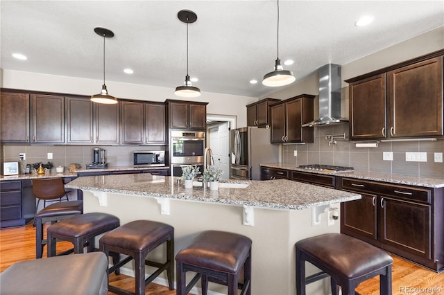 1852 E Sea Drift Dr, Windsor, CO 80550 - kitchen featuring appliances with stainless steel finishes, wall chimney range hood, decorative backsplash, dark brown cabinets, and light wood-style flooring