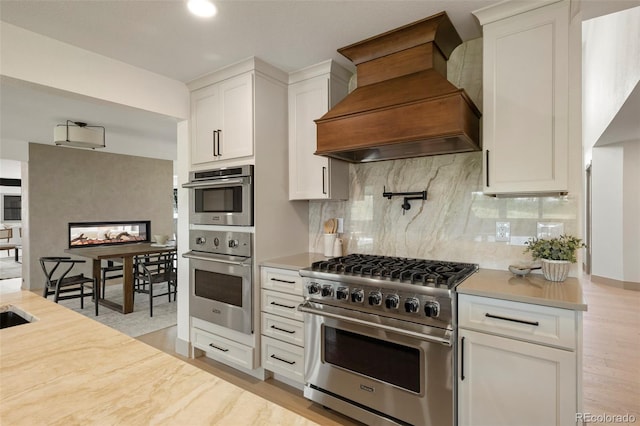 kitchen with custom range hood, backsplash, white cabinetry, appliances with stainless steel finishes, and light hardwood / wood-style floors