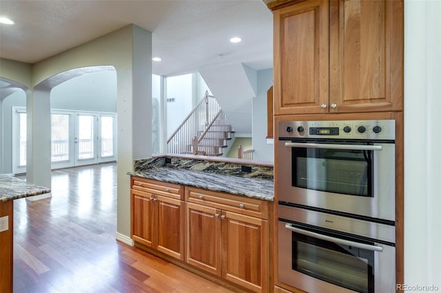 kitchen with double oven, light wood-style flooring, arched walkways, and dark stone counters