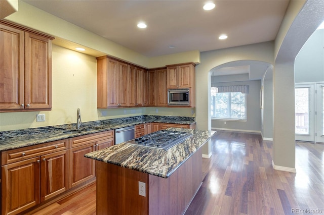 kitchen with light wood finished floors, dark stone counters, arched walkways, stainless steel appliances, and a sink