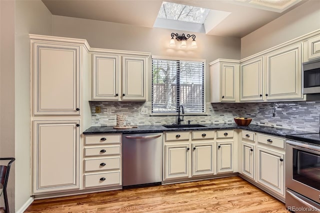 kitchen with cream cabinetry, a sink, a skylight, light wood-style flooring, and appliances with stainless steel finishes