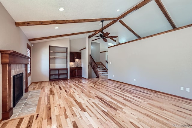 unfurnished living room featuring light wood-type flooring, stairway, a ceiling fan, beamed ceiling, and a tile fireplace