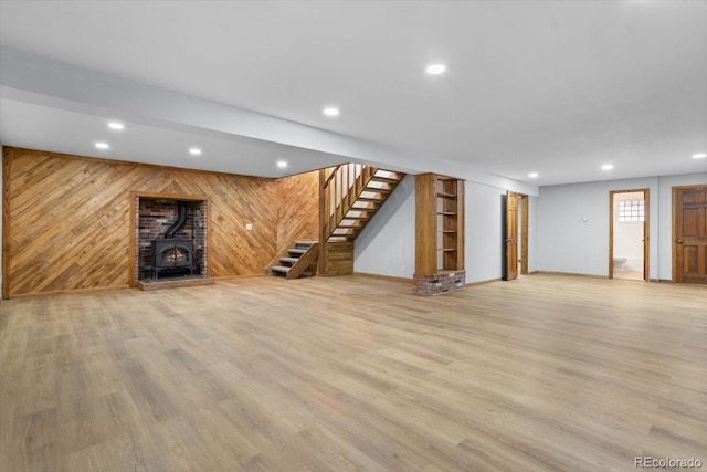 basement with light wood-style flooring, recessed lighting, wooden walls, stairway, and a wood stove