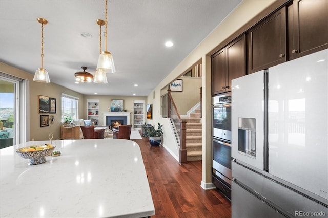 kitchen featuring stainless steel double oven, dark wood-style flooring, a lit fireplace, dark brown cabinets, and refrigerator with ice dispenser