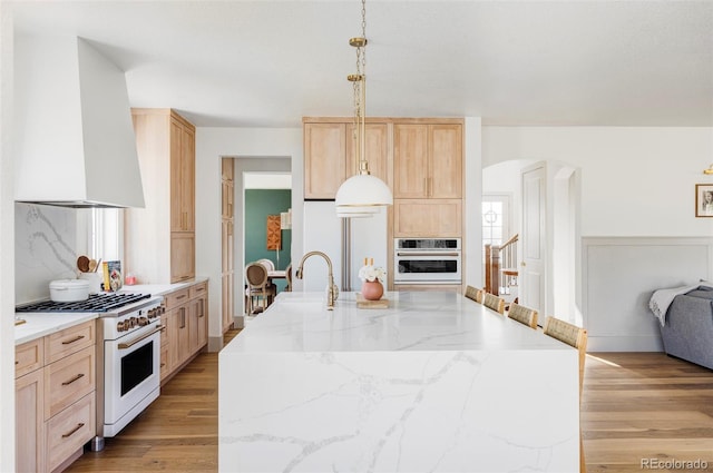 kitchen featuring light brown cabinetry, sink, hanging light fixtures, wall chimney range hood, and white appliances