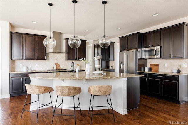 kitchen featuring dark wood-type flooring, wall chimney range hood, a center island with sink, a kitchen breakfast bar, and stainless steel appliances