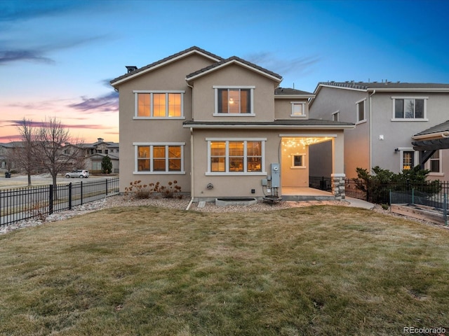 back of house at dusk featuring a lawn, fence, and stucco siding