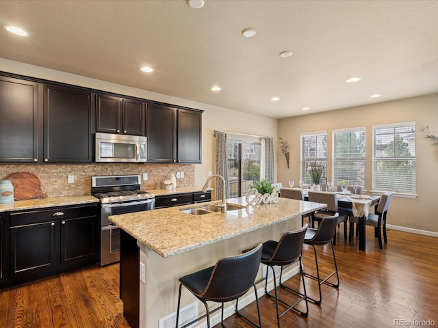 kitchen with a kitchen island with sink, a sink, dark wood-style floors, stainless steel appliances, and decorative backsplash