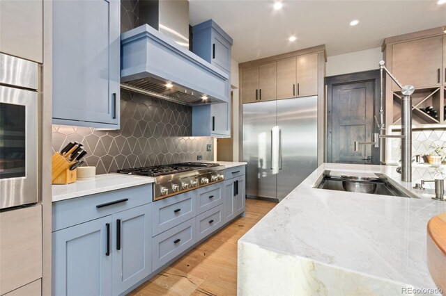3947 Taliesin Way, Fort Collins, CO 80524 - kitchen featuring light stone countertops, light wood-style flooring, stainless steel appliances, decorative backsplash, and wall chimney exhaust hood